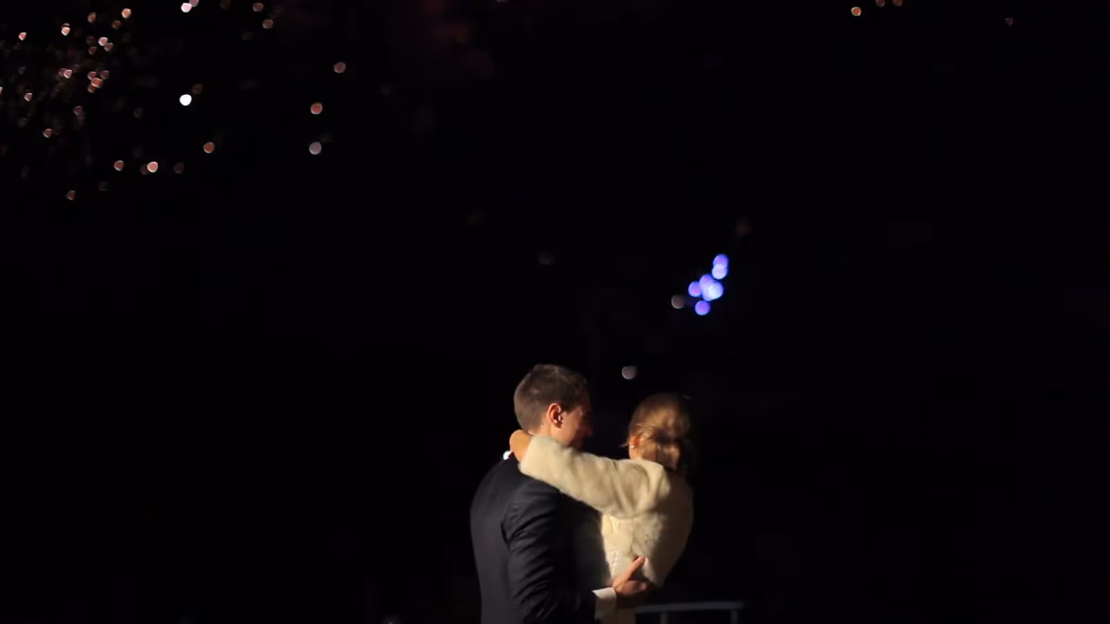 Couple embracing outdoors at night beneath fireworks lighting the dark sky