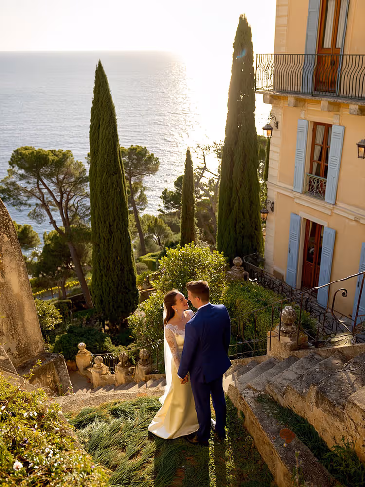 Mariés enlacés sur les marches d'une villa de la Riviera face au coucher de soleil