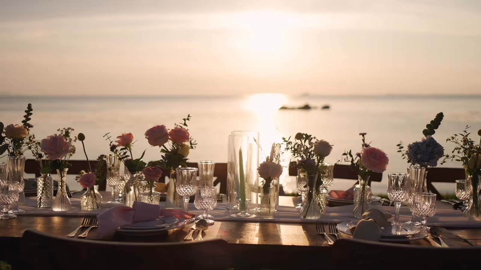 Wooden wedding table set with crystal glasses and pink florals facing the ocean at sunset