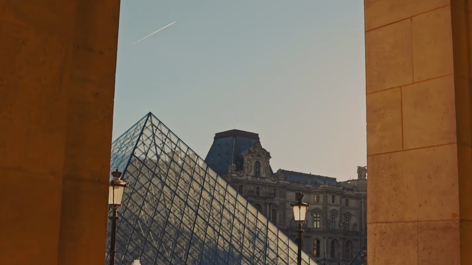 The Louvre glass pyramid and palace facade framed by a stone archway at golden hour