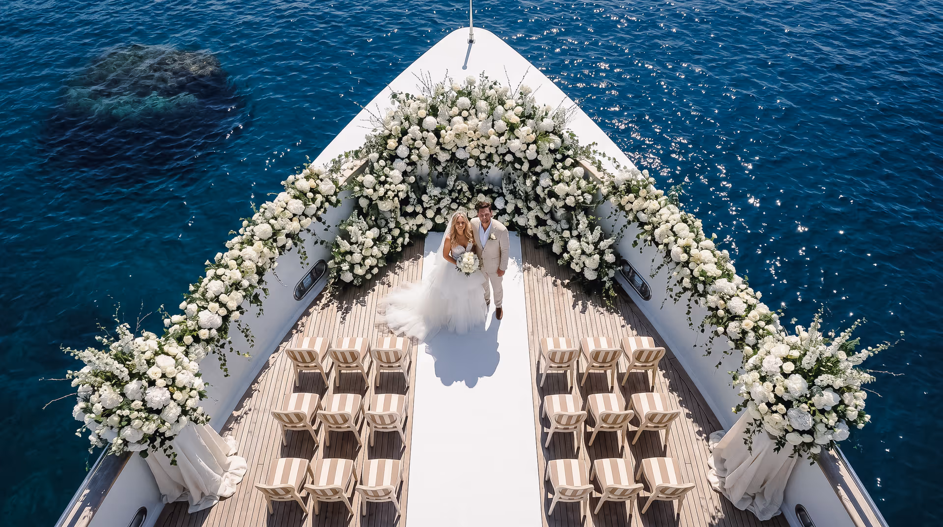 Bride and groom beneath a white floral arch on a yacht bow at sea, ceremony chairs on deck