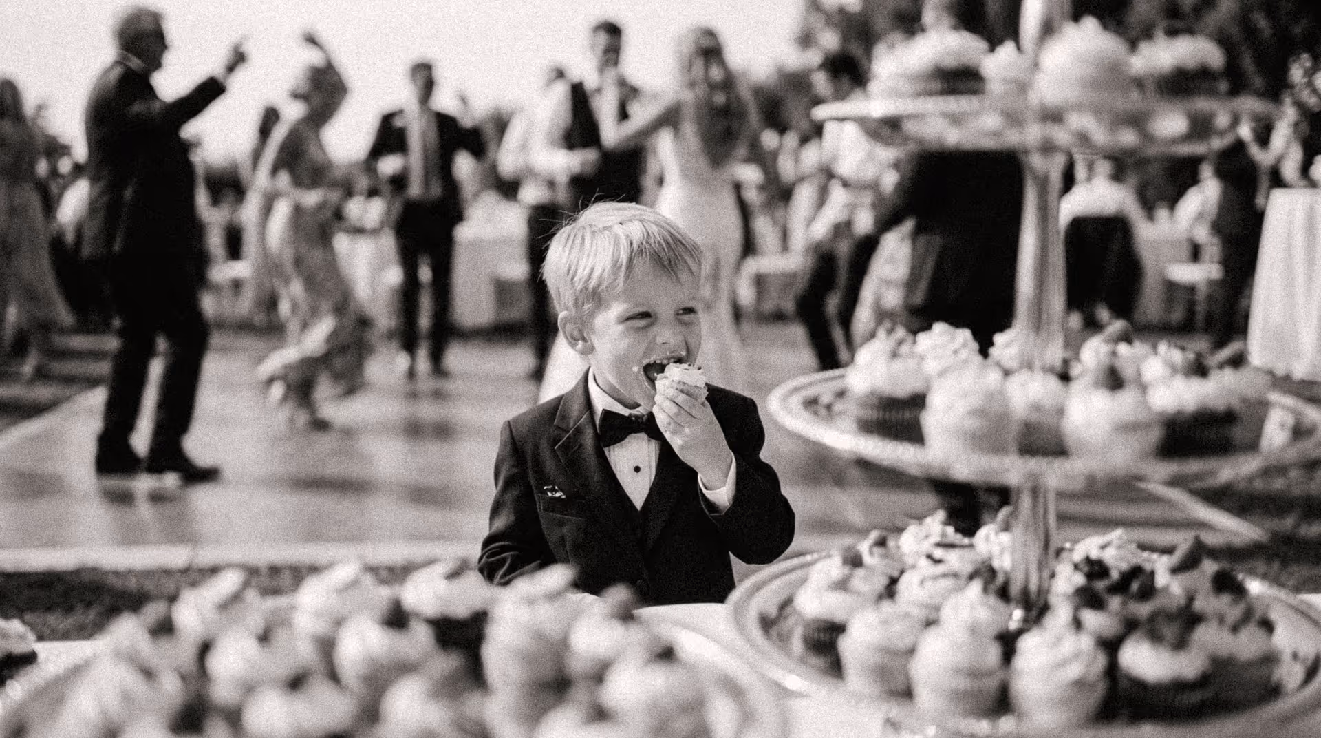 Garçon en smoking goûtant un cupcake au buffet de desserts d'un mariage, invités dansant en arrière-plan