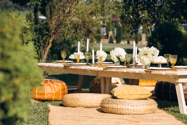 Table basse de jardin avec bougies, hortensias, verres ambrés et poufs tressés sur l'herbe