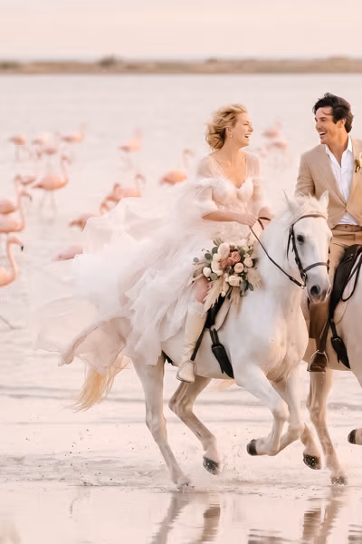 Bride and groom on white Camargue horses galloping through a lagoon, flamingos in the background