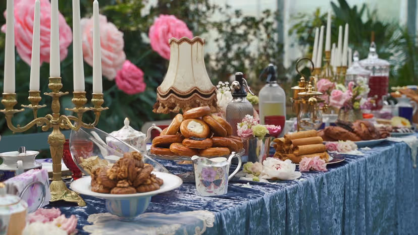 Lavish dessert table with a golden croquembouche, brass candelabra and pink peonies on textured blue cloth.